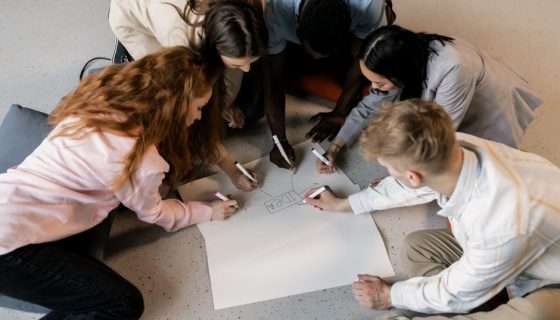 A multicultural team collaborates on an idea using markers and paper on the floor indoors.